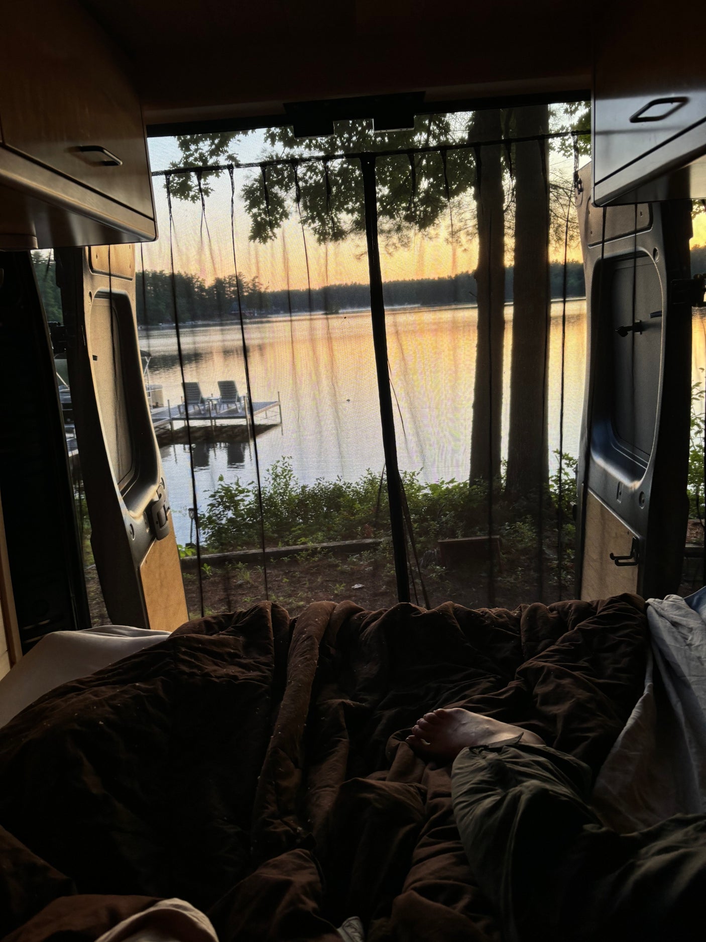 Peaceful rv rental experience: feet propped on a cozy bed, gazing at a stunning lake sunset through a bug screen.