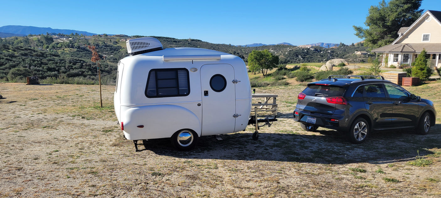 This compact RV rental, towed by a Kia Niro, offers a cozy outdoor escape amidst scenic hills & a distant house.