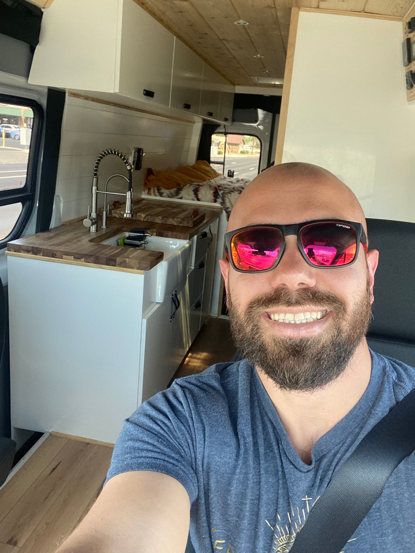 Smiling man in an RV rental camper van, wearing sunglasses. Highlights custom interior with a sink, wooden counter, and cabinets.