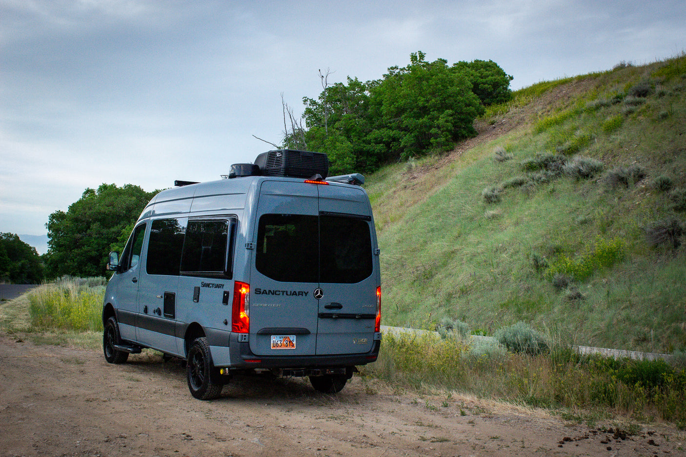Gray Sanctuary Sprinter RV rental on a dirt road, next to a grassy hill. Explore nature in this converted van.