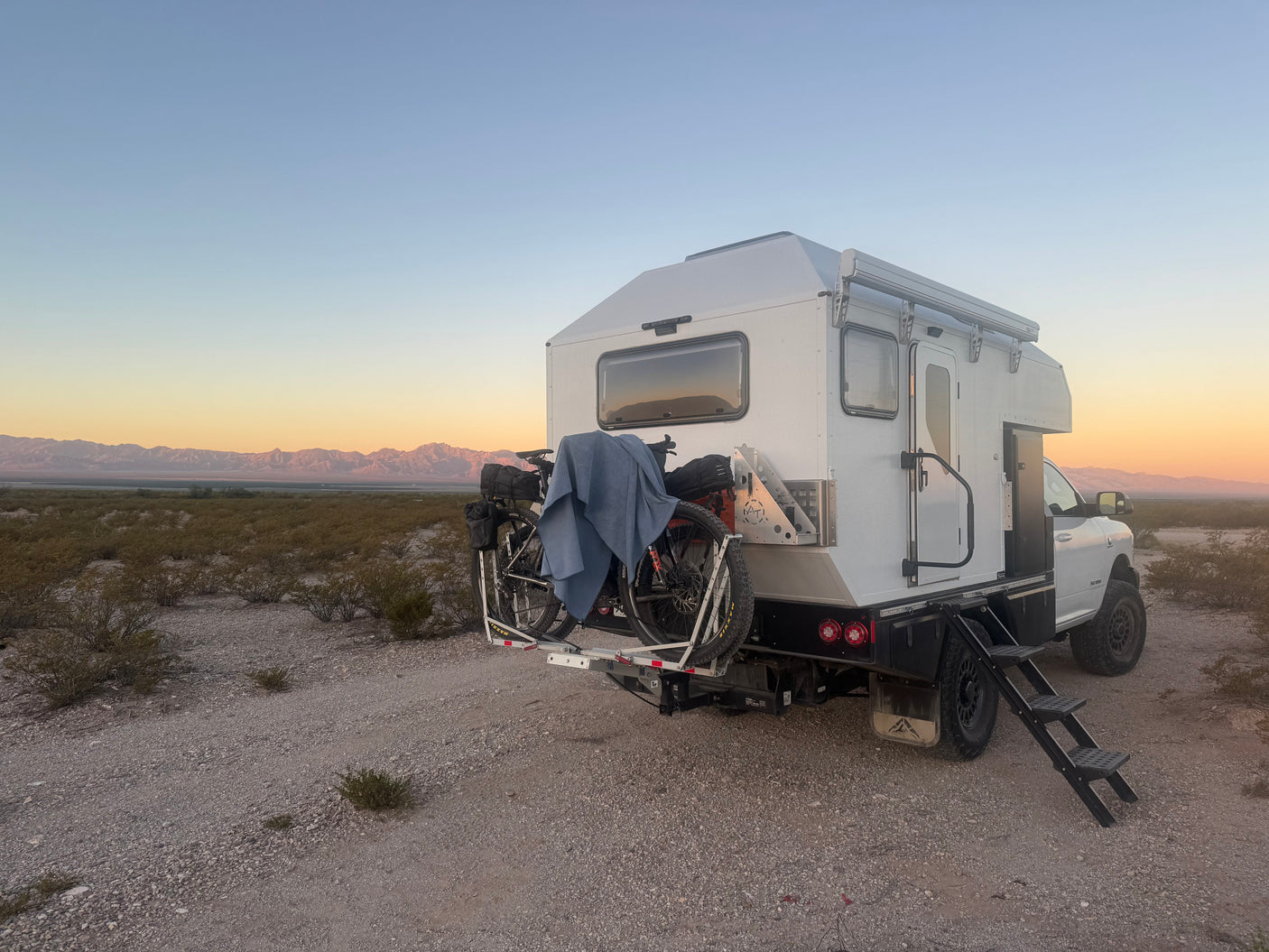 Overland RV rental: white truck camper with bikes and steps, parked on a gravel path in a desert landscape at sunset.