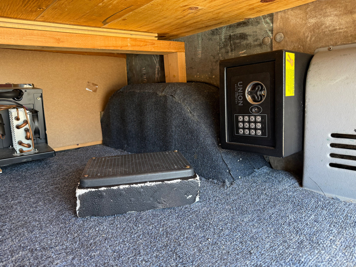 Inside an RV rental: a wall-mounted Union safe with a key warning, a refrigeration unit, and blue carpet on the floor.