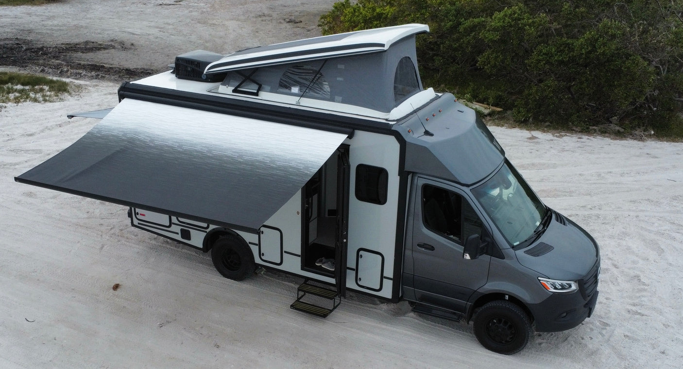 Modern gray rv rental camper van with a deployed pop-up roof tent and extended awning on sandy terrain.