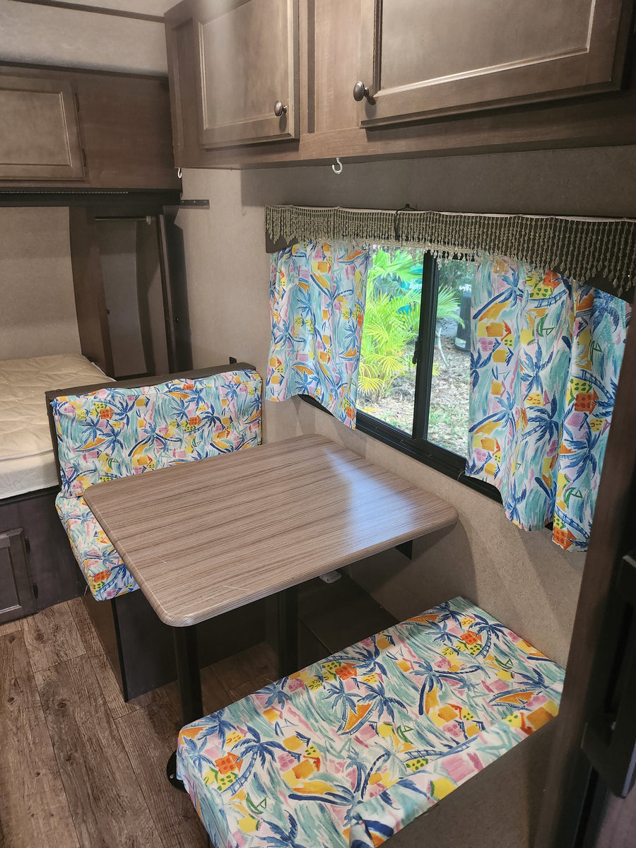 Bright RV rental interior, featuring a dinette with tropical cushions, wood table, window with vibrant curtains, and a nearby bed area.