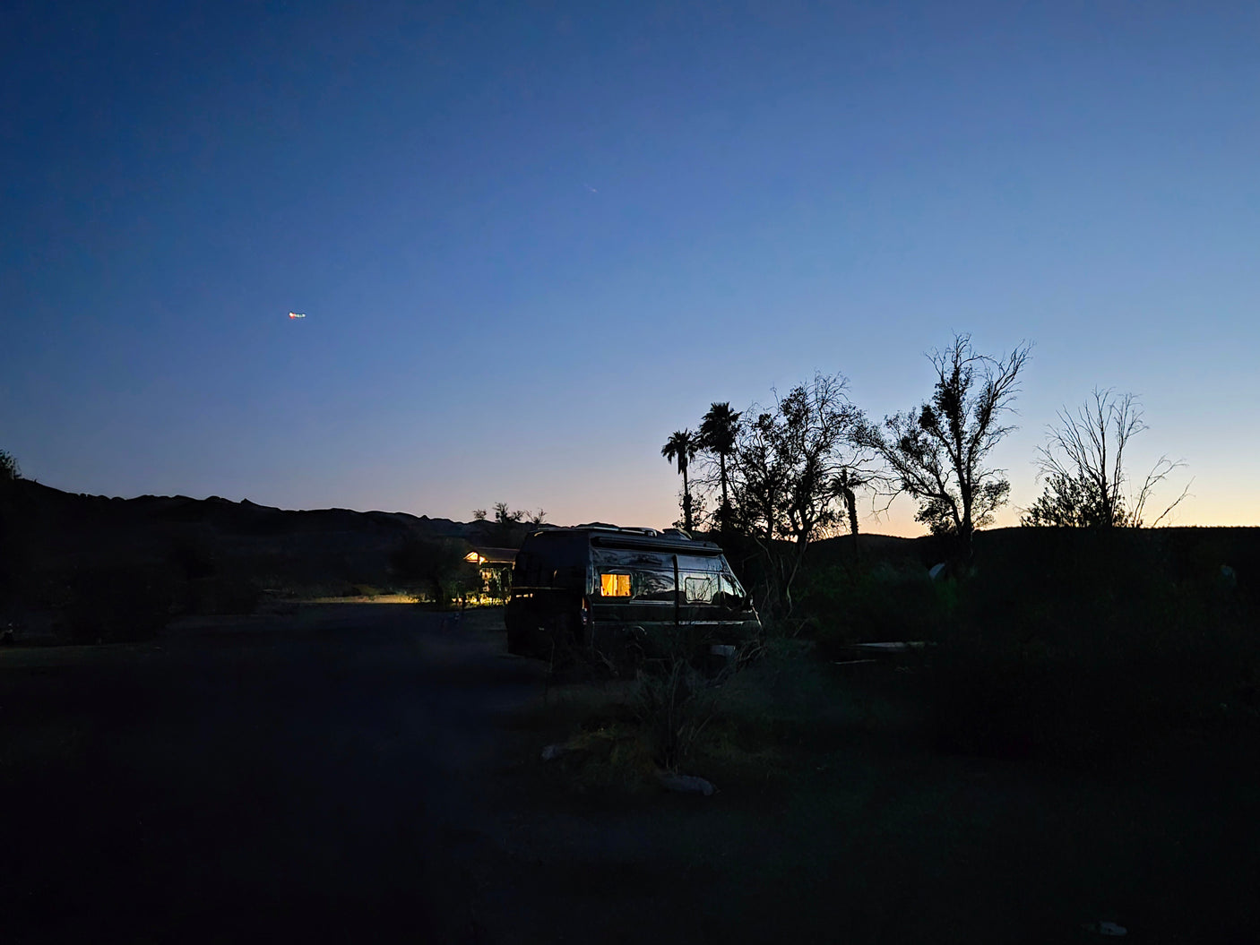 RV rental at twilight. A camper van with glowing windows is parked at a dark campsite, silhouetted by trees & hills.