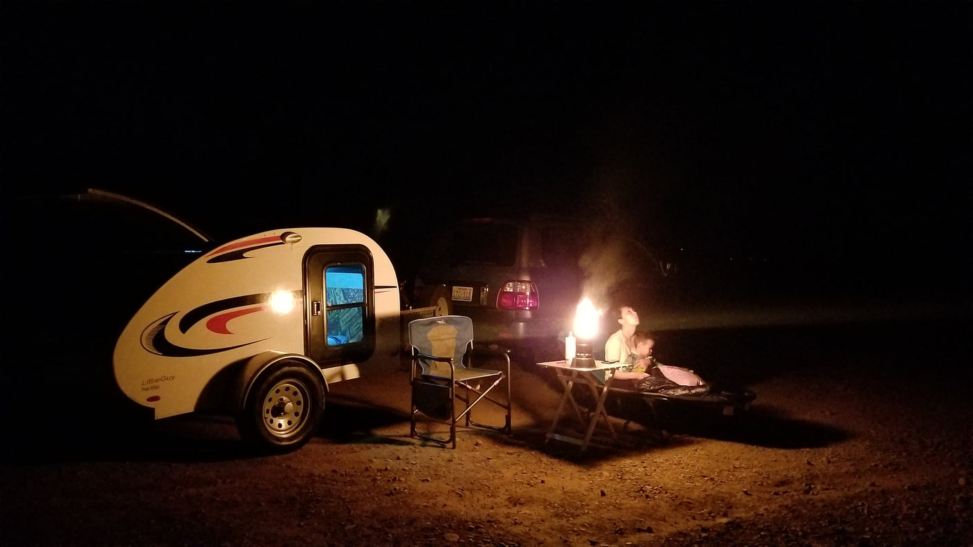 Night view of a camping setup: an **RV rental teardrop trailer**, truck, and person illuminated by a warm **outdoor** light.