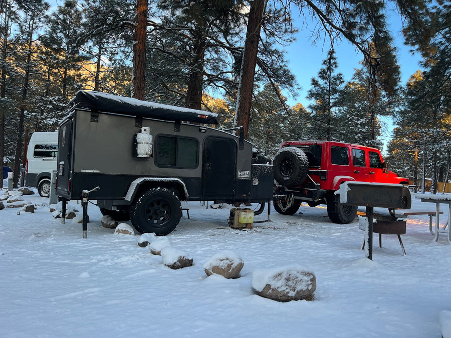 Winter RV rental adventure: A grey overland trailer and red Jeep are parked in a snowy forest campsite