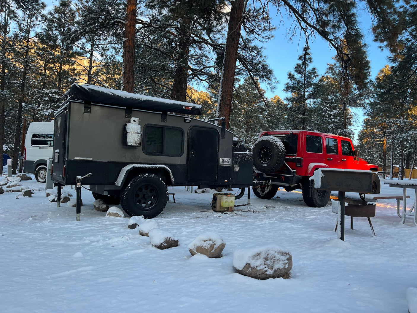Winter RV rental adventure: A grey overland trailer and red Jeep are parked in a snowy forest campsite