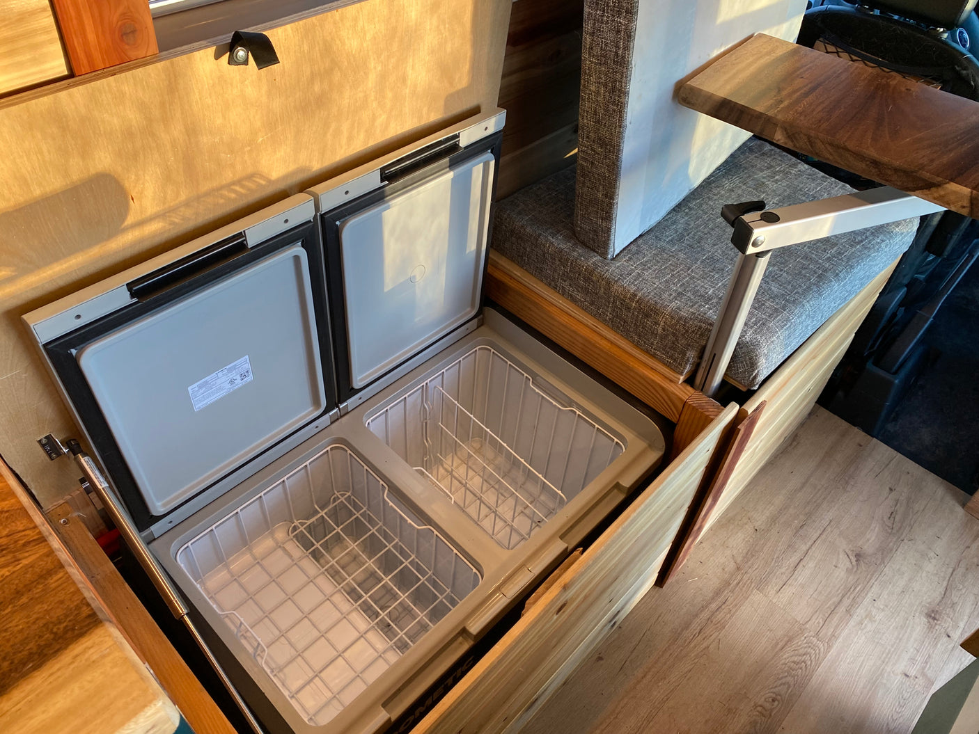 RV rental interior with an open dual-zone portable fridge/freezer in custom wood cabinetry, next to a cushioned bench and adjustable table.