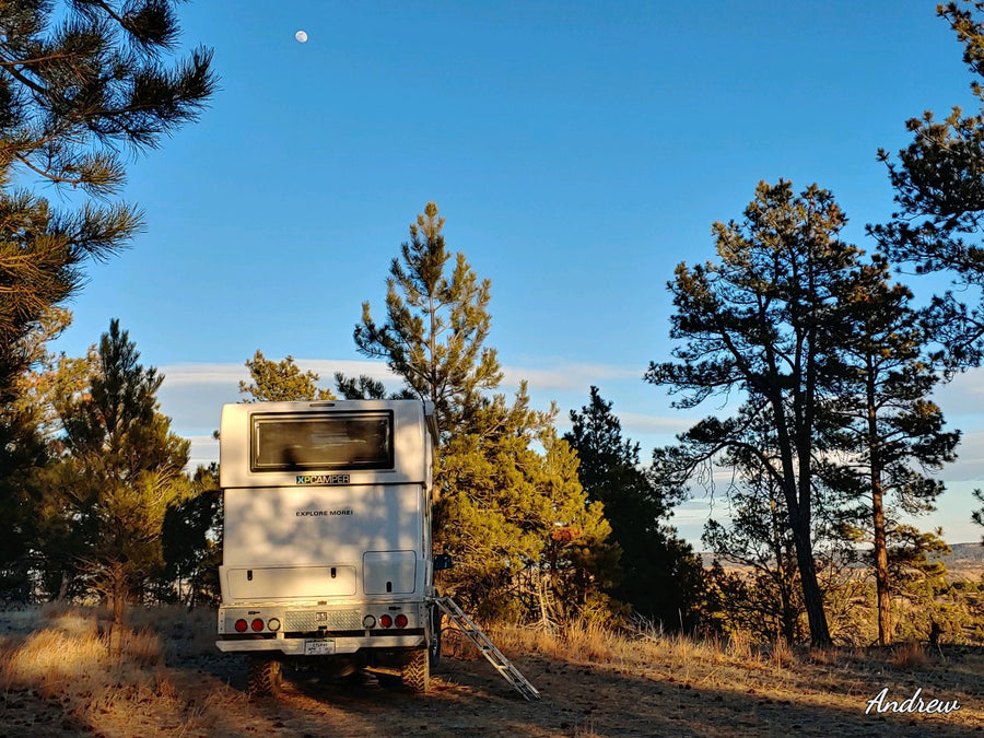 with chairs set up.|Scenic **RV rental** adventure: White camper parked among pines under a bright blue