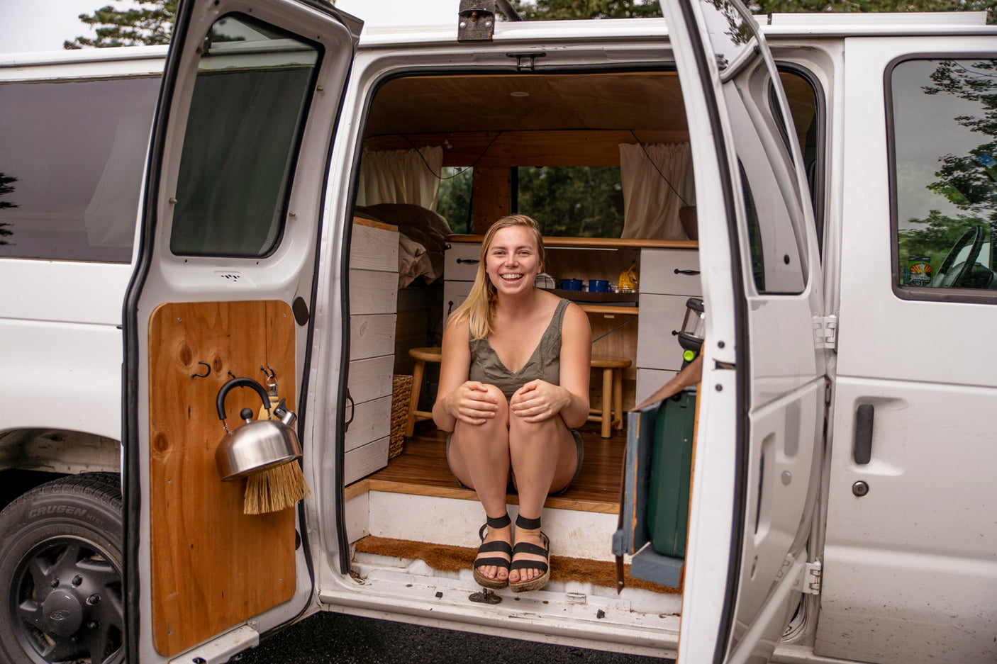 A smiling blonde woman sits inside a white converted camper van with open doors, showcasing its cozy wood-paneled interior. Perfect for an RV rental adventure.