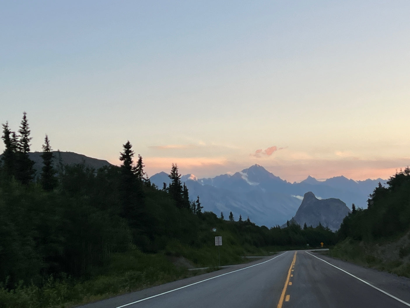 Scenic road ahead towards impressive mountains with a sunset sky. Dense greenery flanks the path, perfect for an RV rental journey.