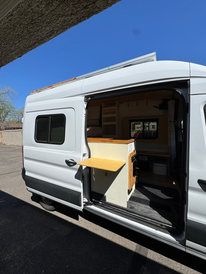 A white custom rv rental van with its sliding door open, revealing a cozy interior with a bed, shelves, and pull-out table.