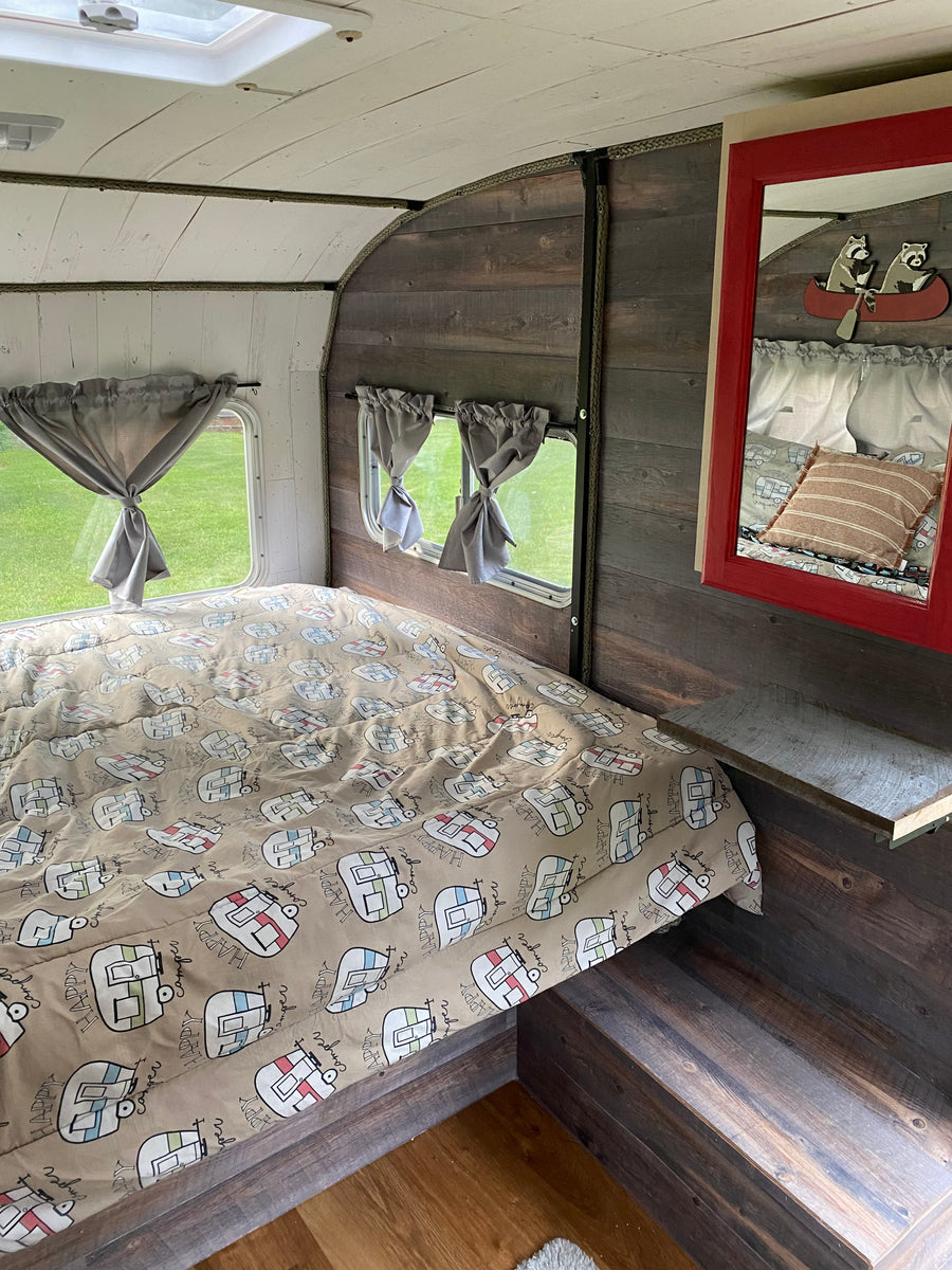Relax in this cozy rv rental bedroom, featuring a "Happy Camper" comforter, rustic wood walls, and bright windows.