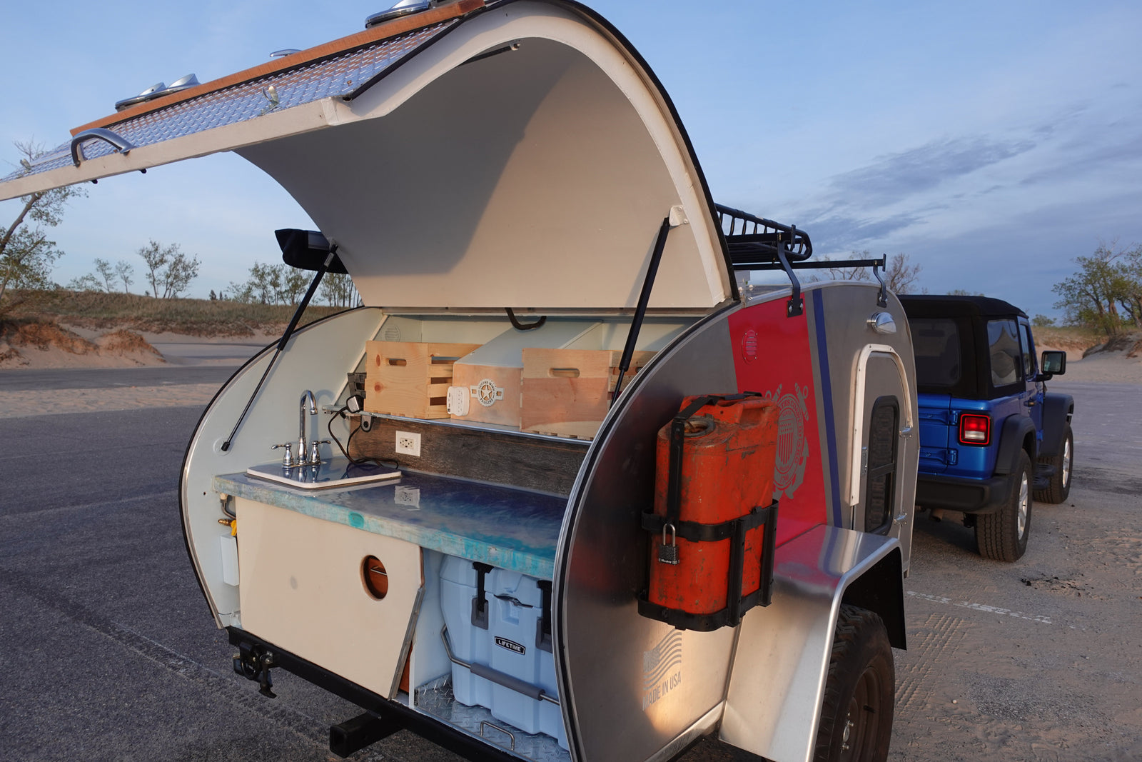Discover this rv rental teardrop camper's kitchenette with sink, counter, and cooler. Parked by a Jeep in sandy terrain.