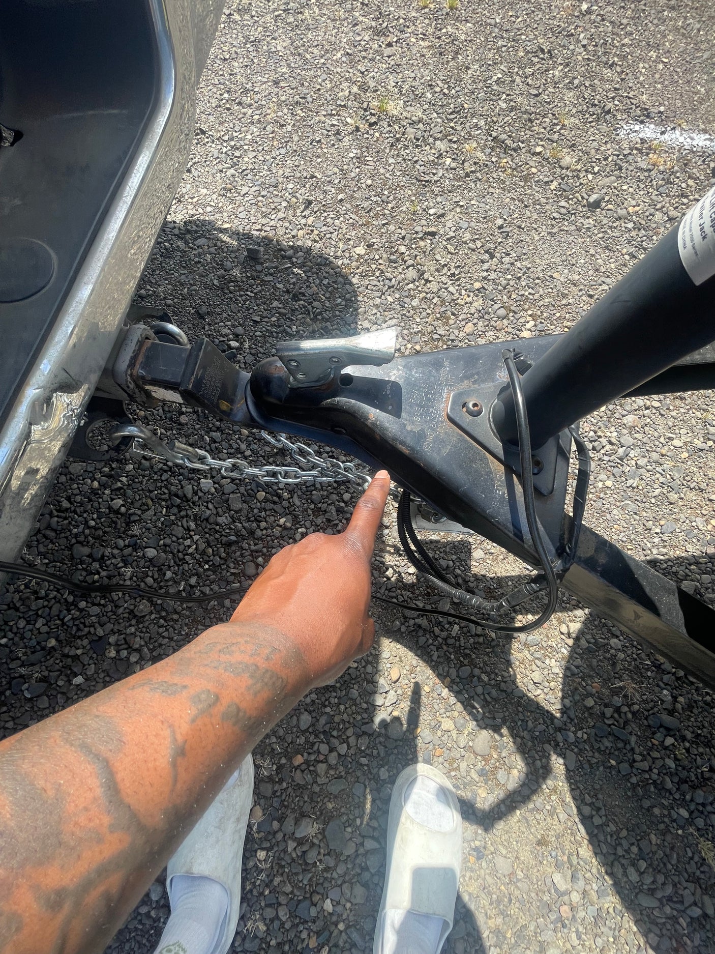 A person points to the RV rental's trailer hitch, showing the secure connection and safety chains on a gravel surface.