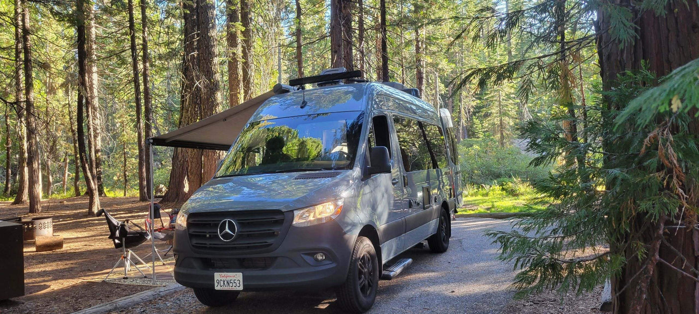 Perfect RV rental for a forest getaway. Gray camper van under an awning at a sunny campsite amidst tall trees.