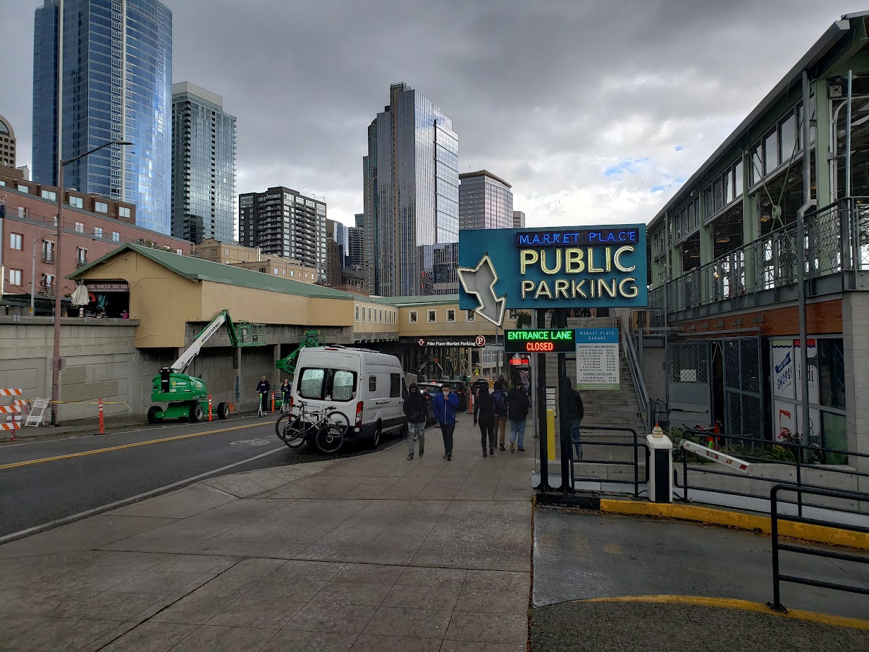 Urban view: white RV rental van with bikes on a city street. 'Public Parking' sign (entrance closed). Tall buildings & pedestrians are seen.