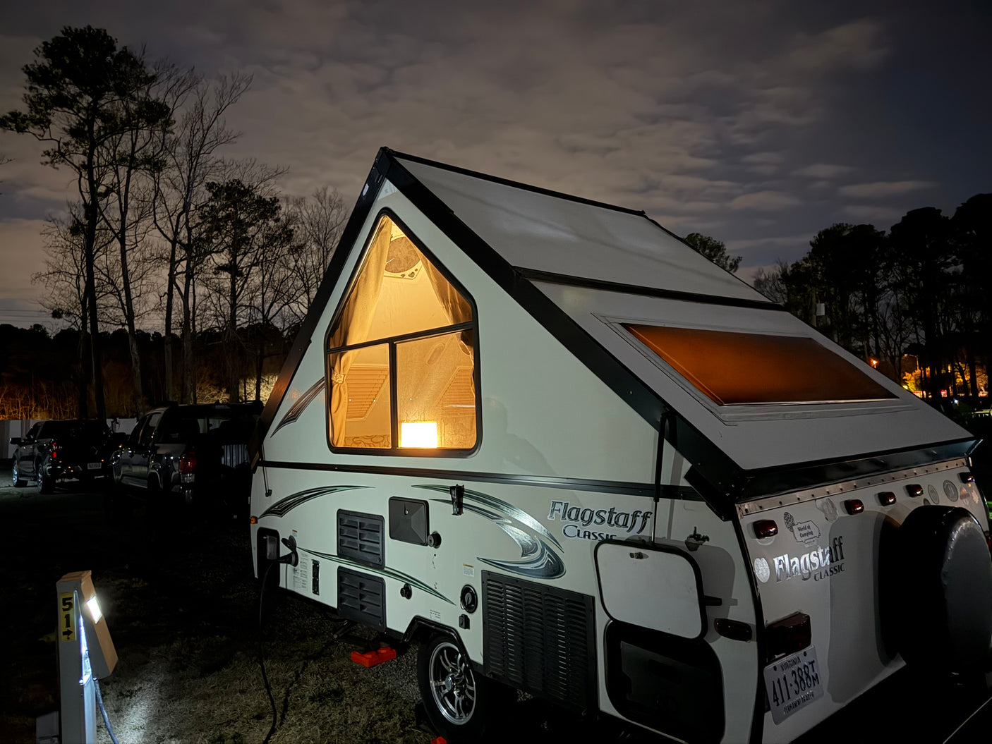 An A-frame rv rental (Flagstaff Classic) glows warmly at night in a campsite. Trees and cloudy sky above.