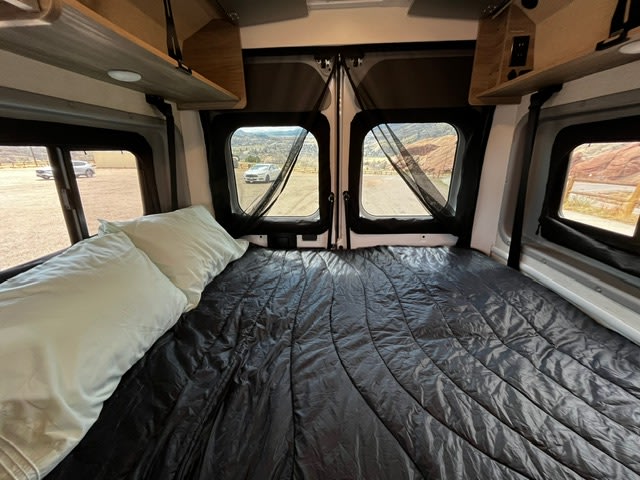 This RV rental shows a comfy bed with a dark comforter and light pillows, offering a scenic outdoor view.