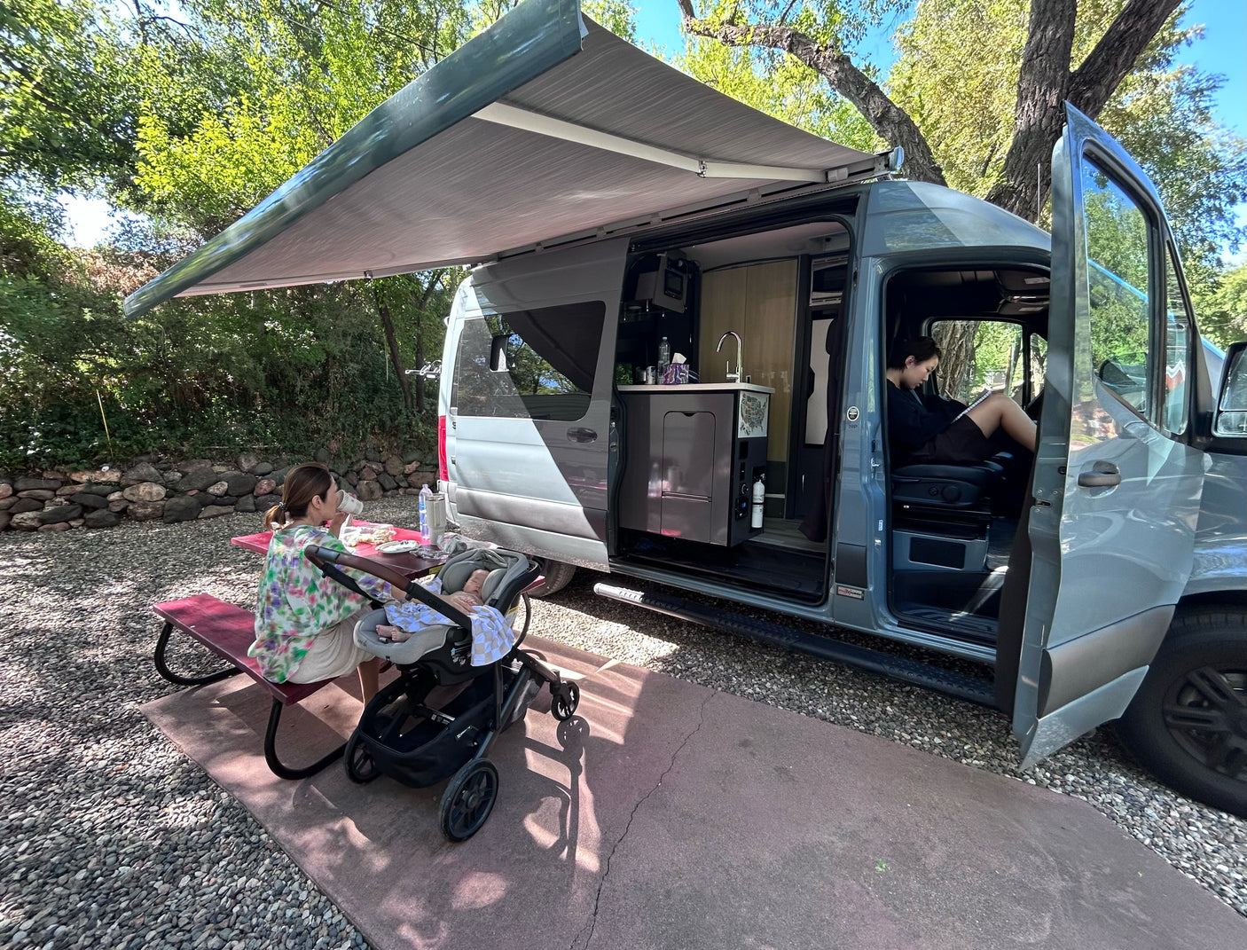 An RV rental vacation: two women and a baby relax outdoors by their camper van, one at a picnic table, one in the driver's seat.
