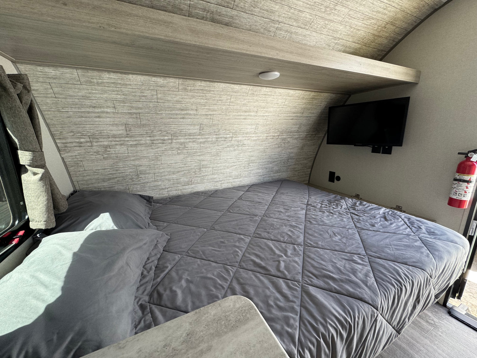 Cozy RV rental bedroom featuring a grey bed, overhead shelf, and wall-mounted TV. Perfect for road trip comfort.