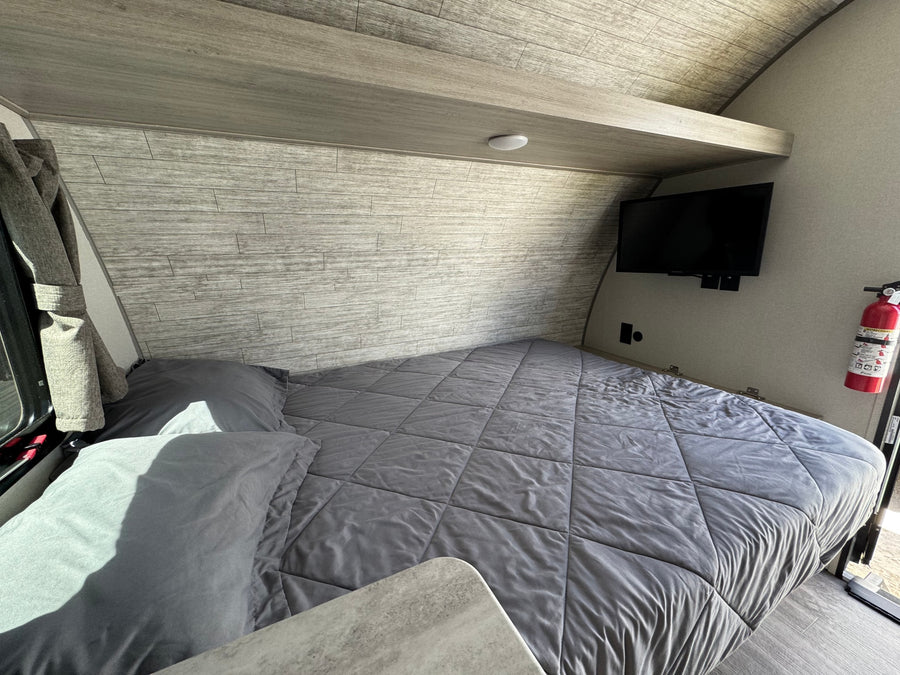 Cozy RV rental bedroom featuring a grey bed, overhead shelf, and wall-mounted TV. Perfect for road trip comfort.