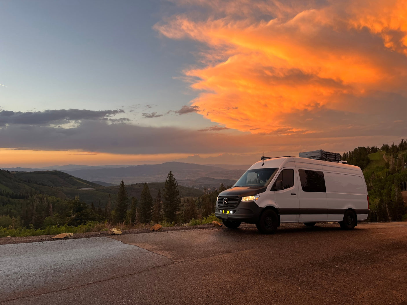White Mercedes RV rental van at a mountain overlook. Vibrant orange sunset clouds fill the sky above scenic valleys.