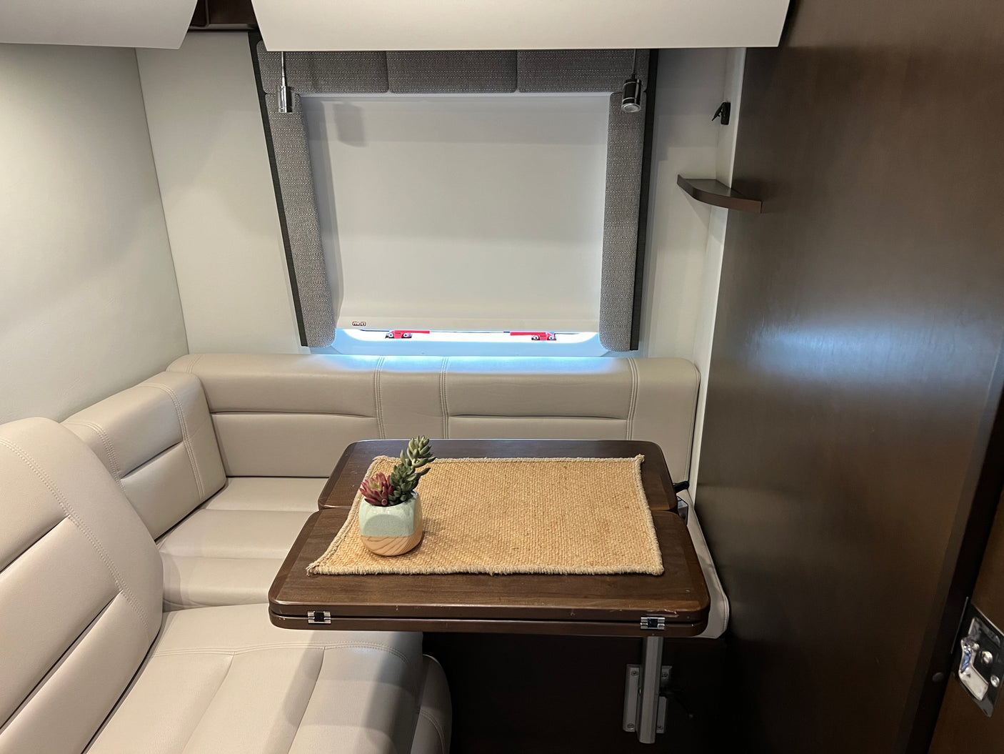 This RV rental features a comfortable dinette: plush seating, wooden table with decor, and adjustable window blinds.