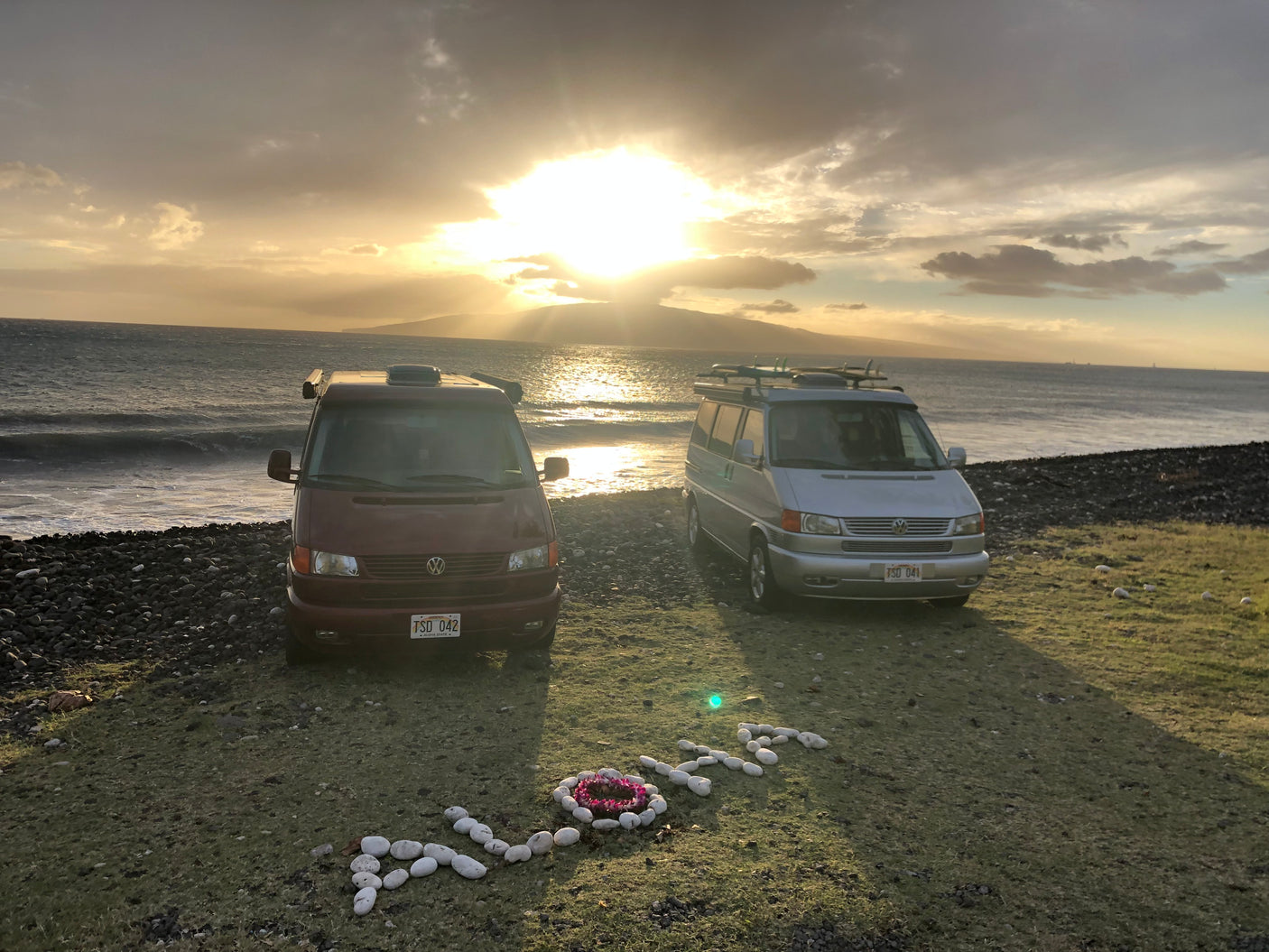 Two RV rental camper vans are parked on a grassy beach in Hawaii at sunset, with 'Aloha' spelled out in white rocks.
