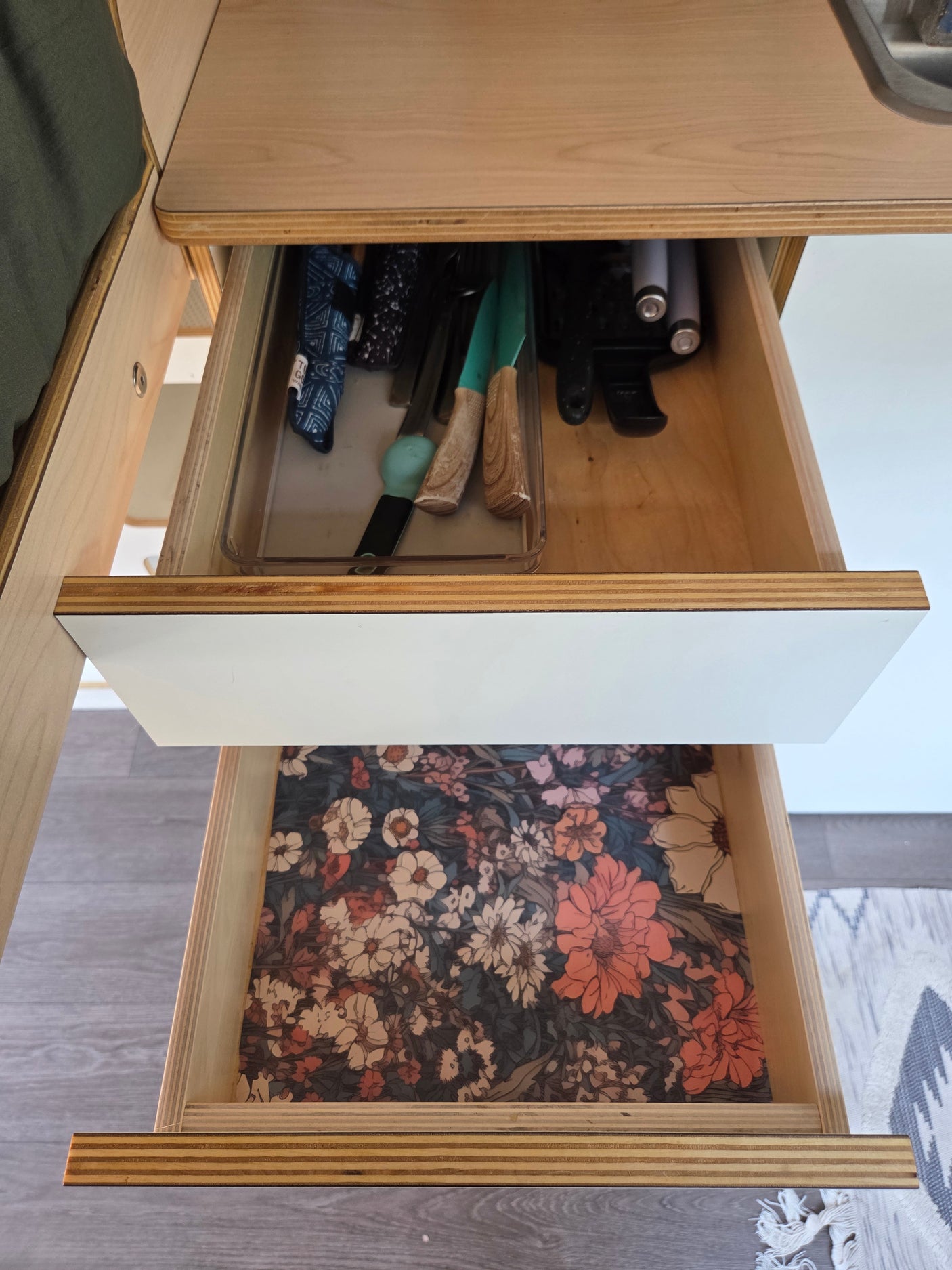 RV rental kitchen drawers: top drawer holds utensils in an organizer, bottom drawer features a vibrant floral liner. Plywood design.