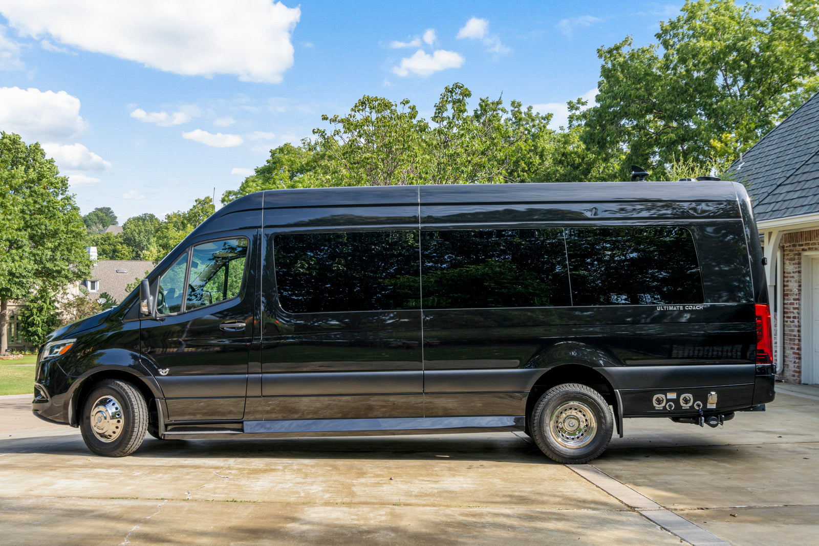 This black luxury Mercedes-Benz Sprinter van, an ideal RV rental, is parked on a sunny driveway. It features sleek tinted windows.