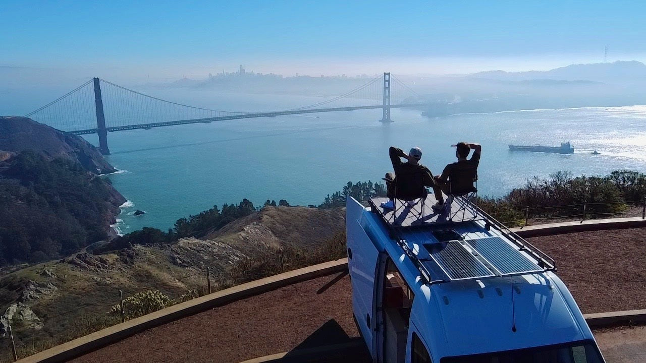 Golden Gate Bridge view from an RV rental. Two people relax on van's rooftop at a scenic overlook.