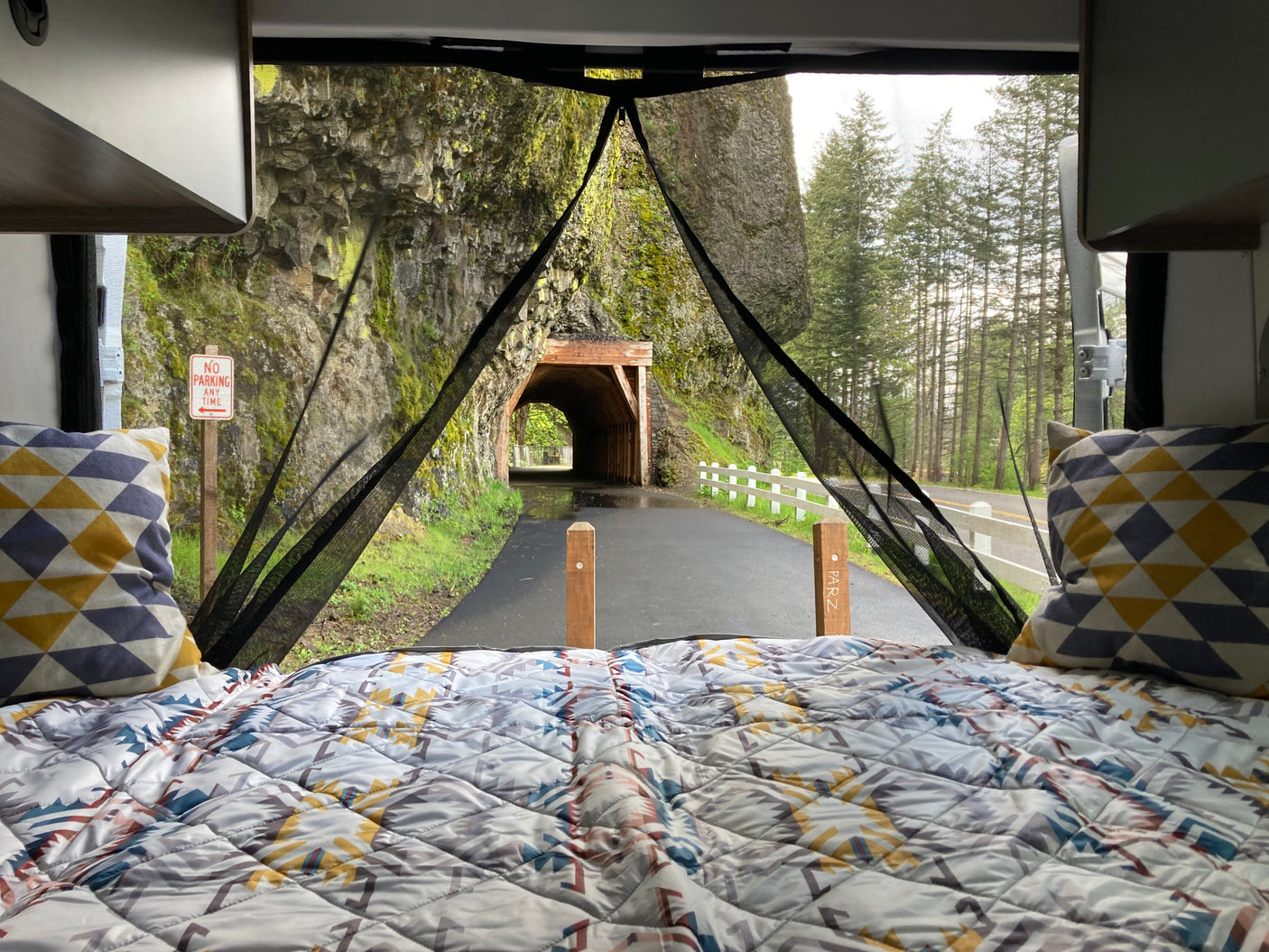 Scenic RV rental view from inside: Bed with patterned quilt, looking out to a tunnel entrance, path, and tall trees.