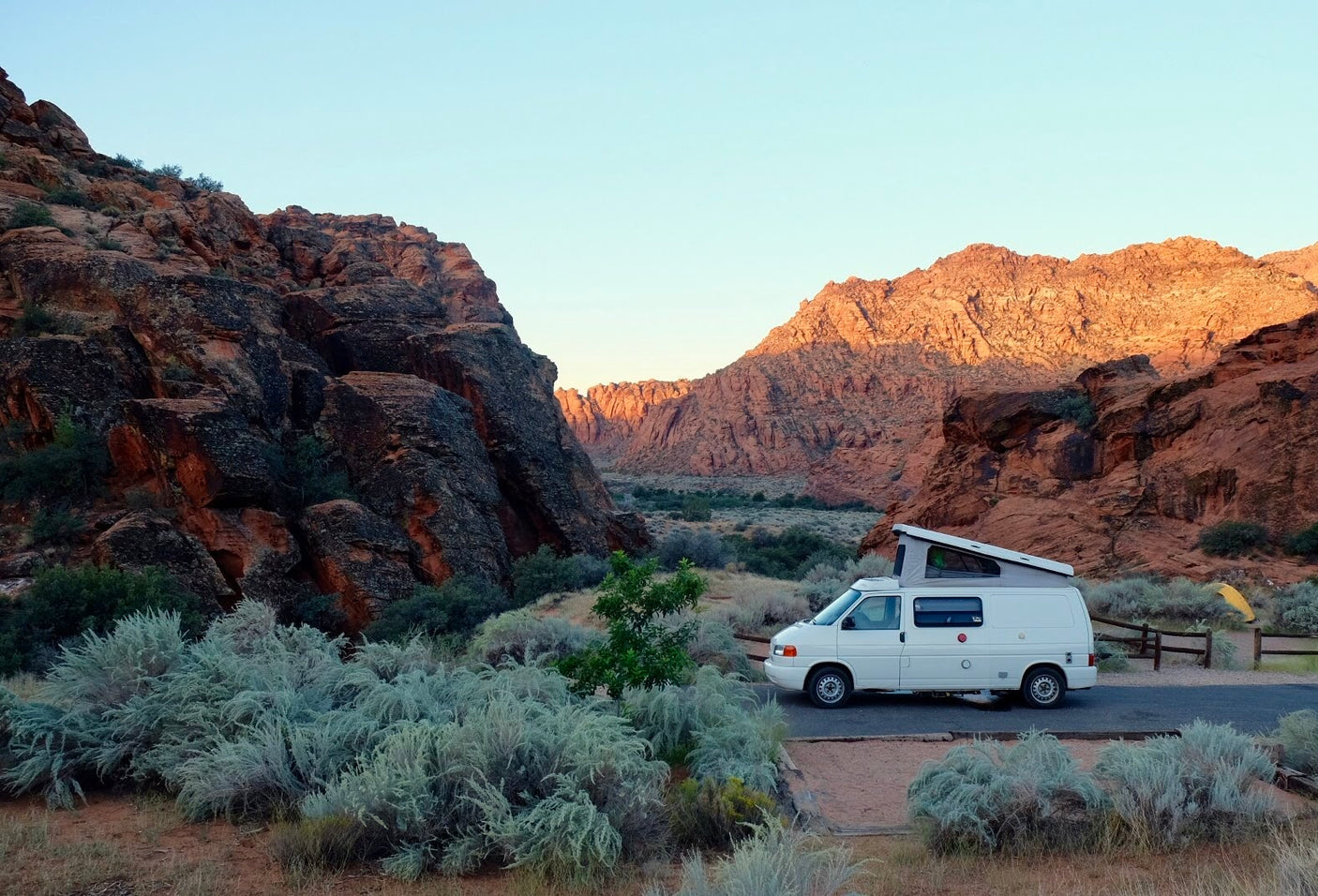 Explore stunning desert landscapes with an RV rental! A white camper van with a pop-top is parked among red rock cliffs and desert plants.