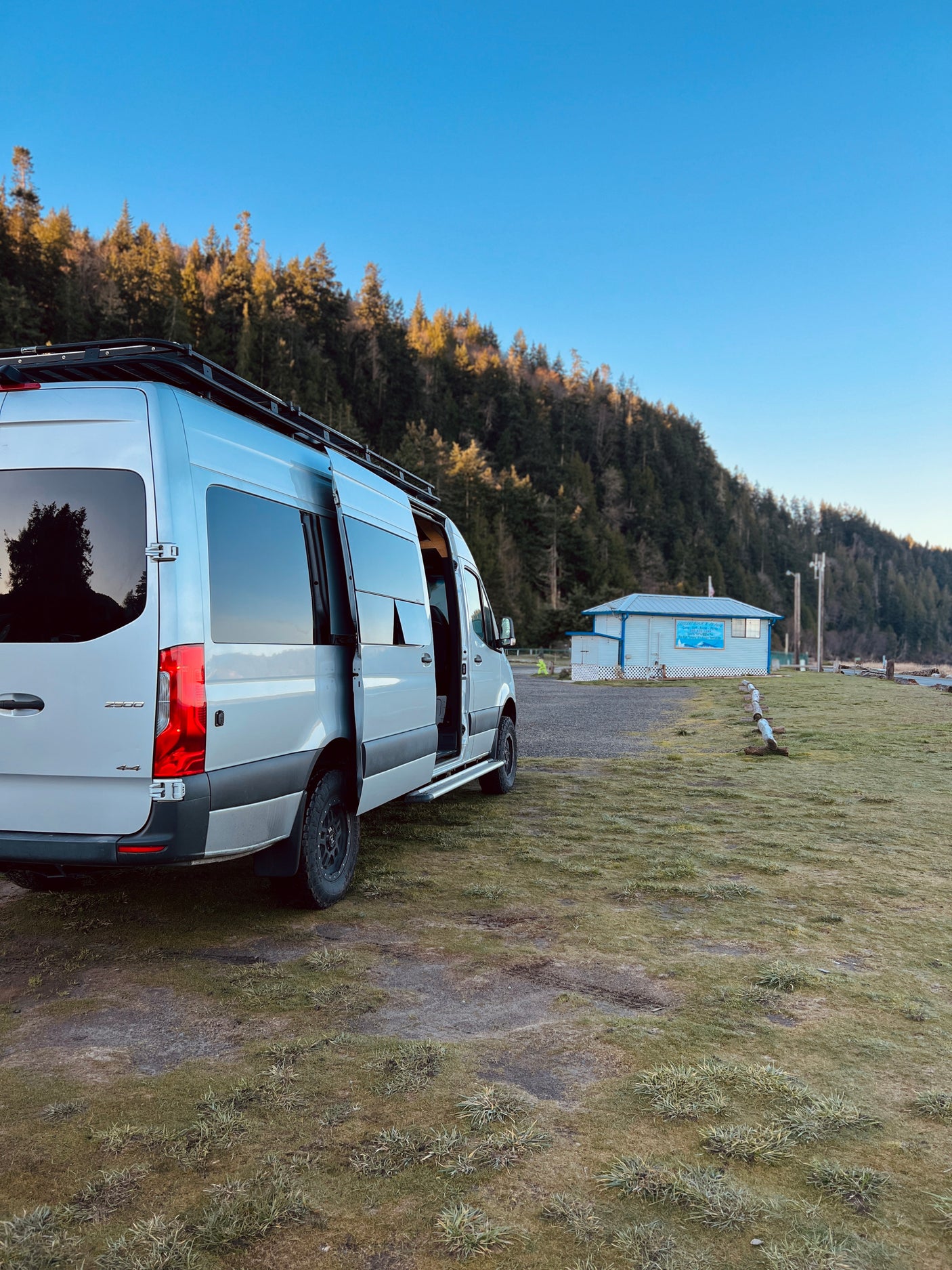 Silver Sprinter camper van (rv rental) with roof rack and open door, parked by a forest and 'Ocean Park Camp & Kayak' building.
