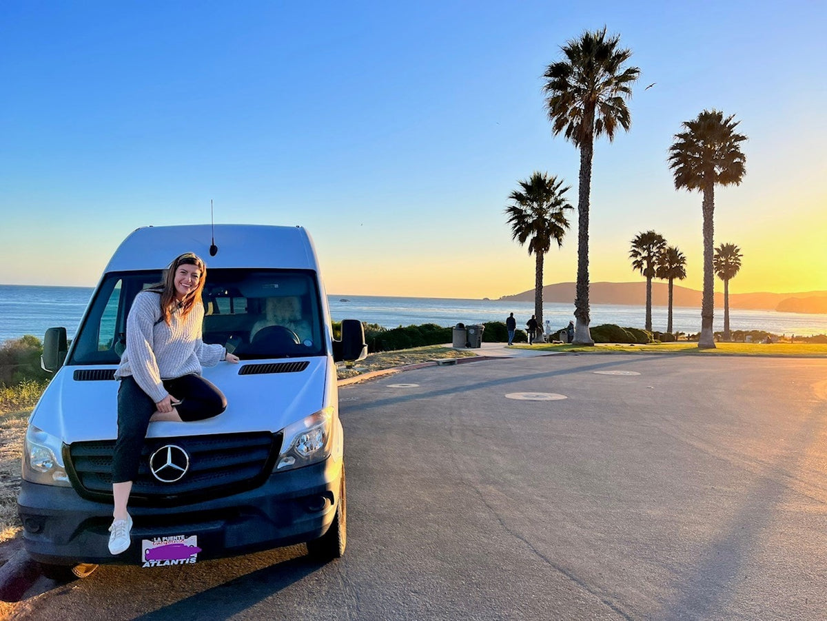 Coastal rv rental adventure! Woman on white van at sunset, overlooking ocean and palm trees.