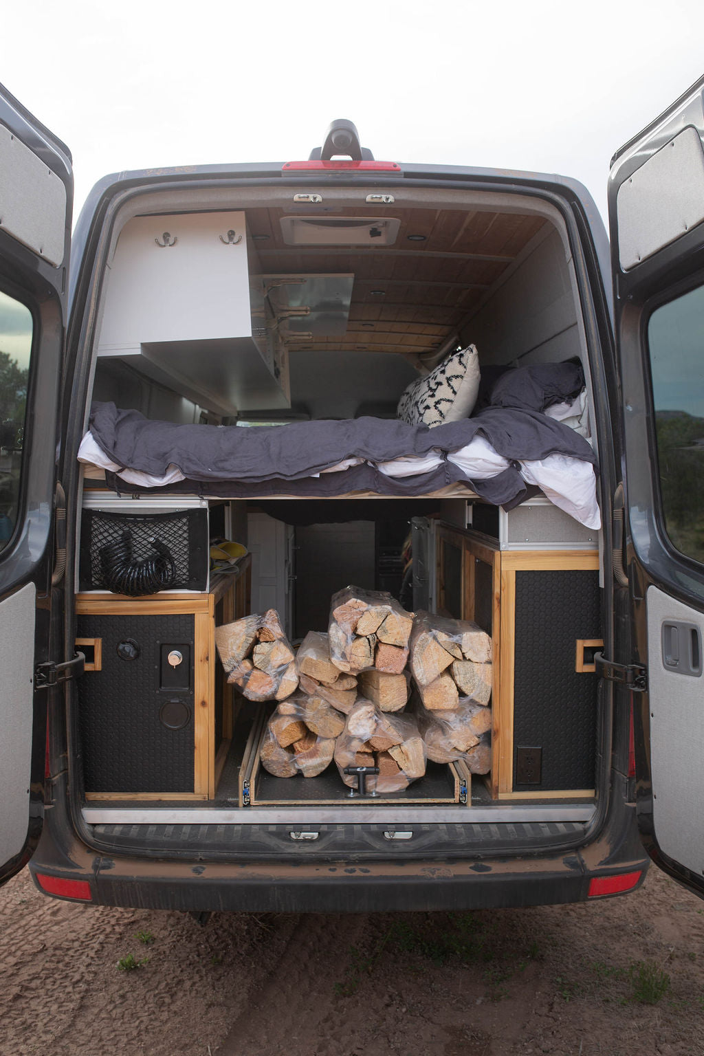 RV rental camper van interior: elevated bed, storage, and firewood on pull-out trays at the rear doors.