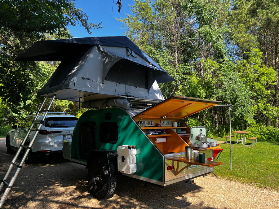 RV rental adventure: SUV, green teardrop trailer with outdoor kitchen, & rooftop tent deployed for camping in sunny woods.
