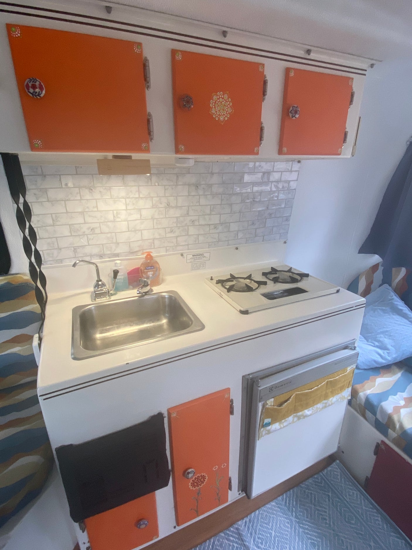 Cozy RV rental kitchen with vibrant orange cabinets, subway tile backsplash, sink, stove, and compact fridge. Perfect for road trips.