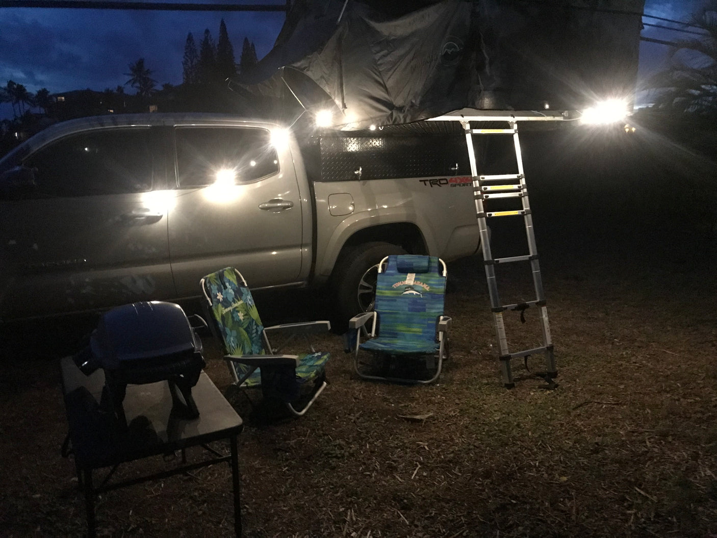 Night scene with a pickup truck, its rooftop tent and ladder set up for camping. Two chairs and a grill are ready for an RV rental trip.