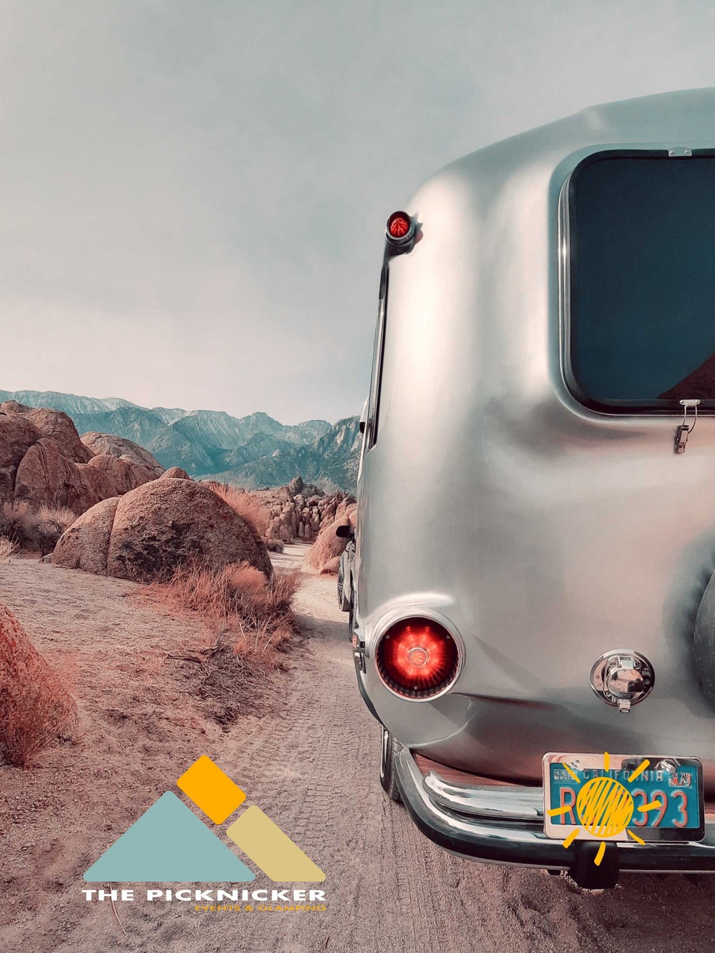 A shiny silver Airstream RV rental on a dirt road, amidst rugged desert rocks and distant mountains, ideal for adventure.