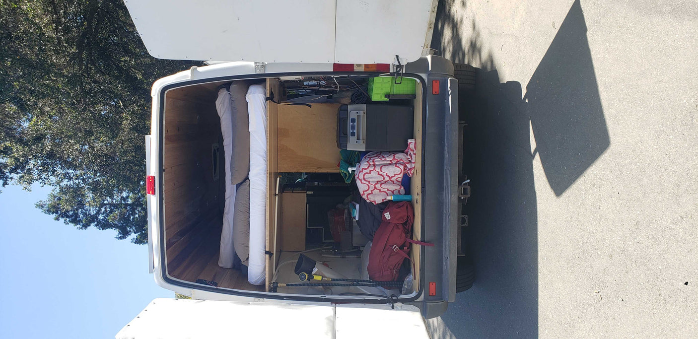 An RV rental camper van's open rear doors reveal a well-organized interior with rolled bedding, a portable fridge, and storage.
