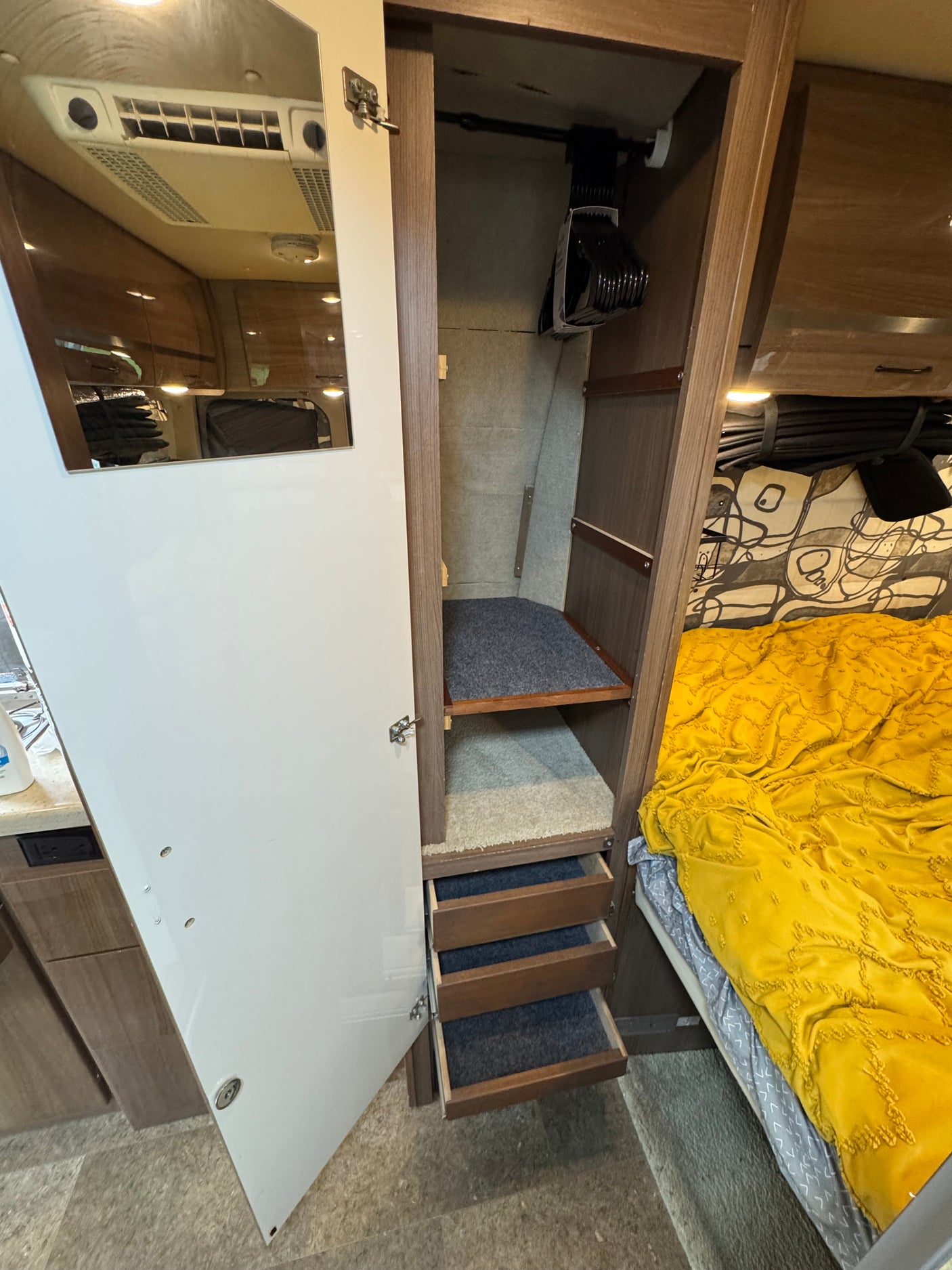 RV rental storage: Open closet with hanging rod, shelves, and carpet-lined drawers. A bed with a yellow blanket is visible nearby.
