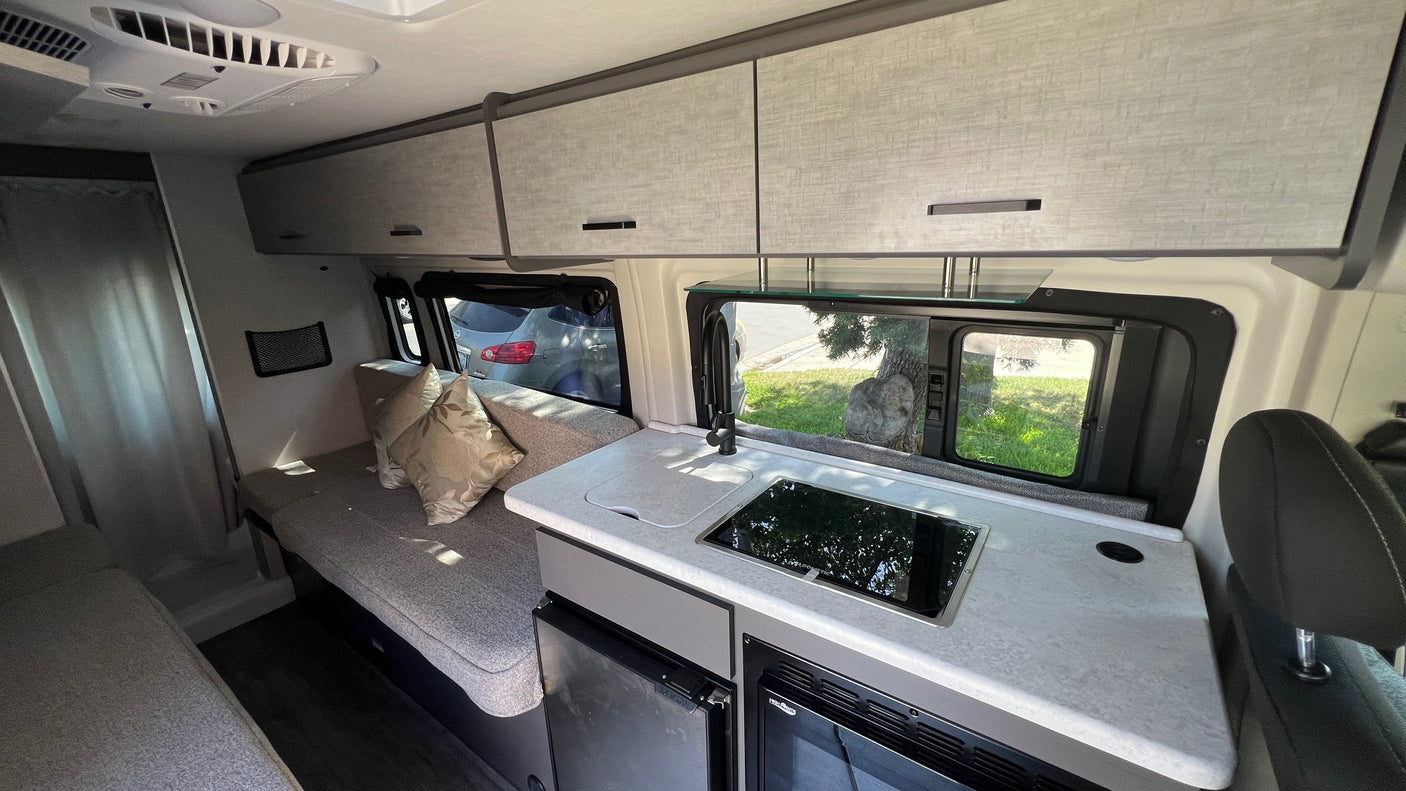 Inside an RV rental: Compact kitchen with sink & cooktop, overhead storage, and lounge seating. Ideal for travel.