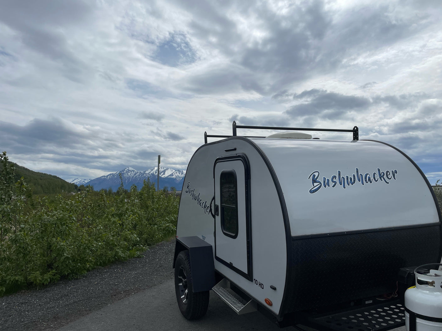 Bushwhacker teardrop camper RV rental parked with snowy mountains and cloudy skies in background.