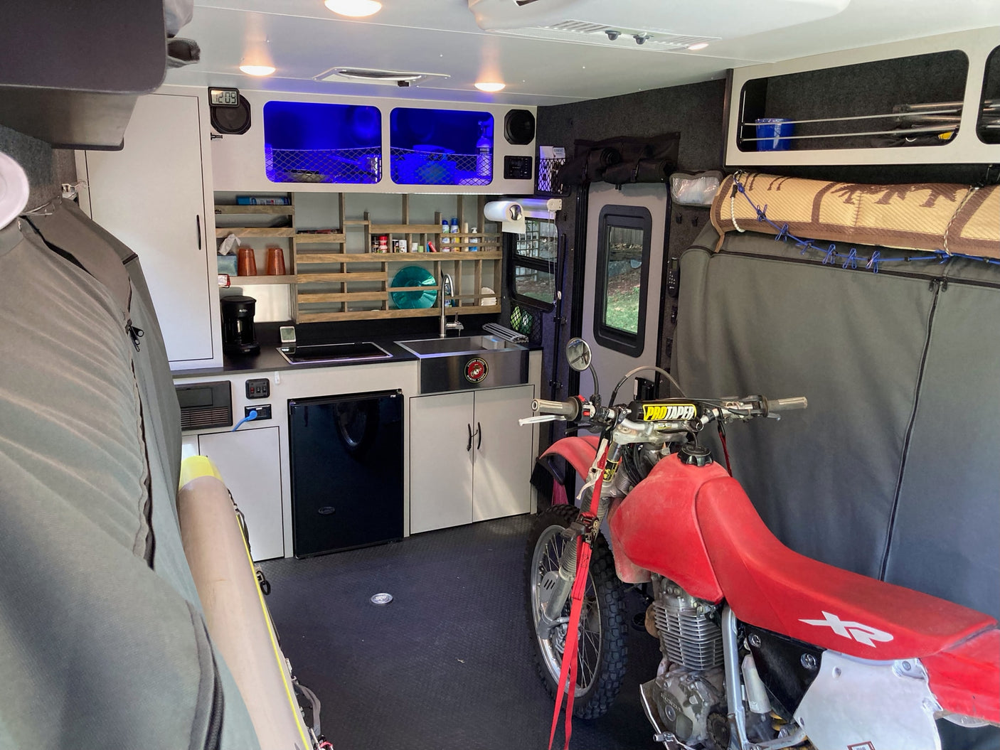 Interior of an rv rental toy hauler, featuring a dirt bike secured alongside a compact kitchen, storage, and ambient blue lighting.