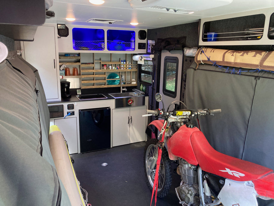 Interior of an rv rental toy hauler, featuring a dirt bike secured alongside a compact kitchen, storage, and ambient blue lighting.
