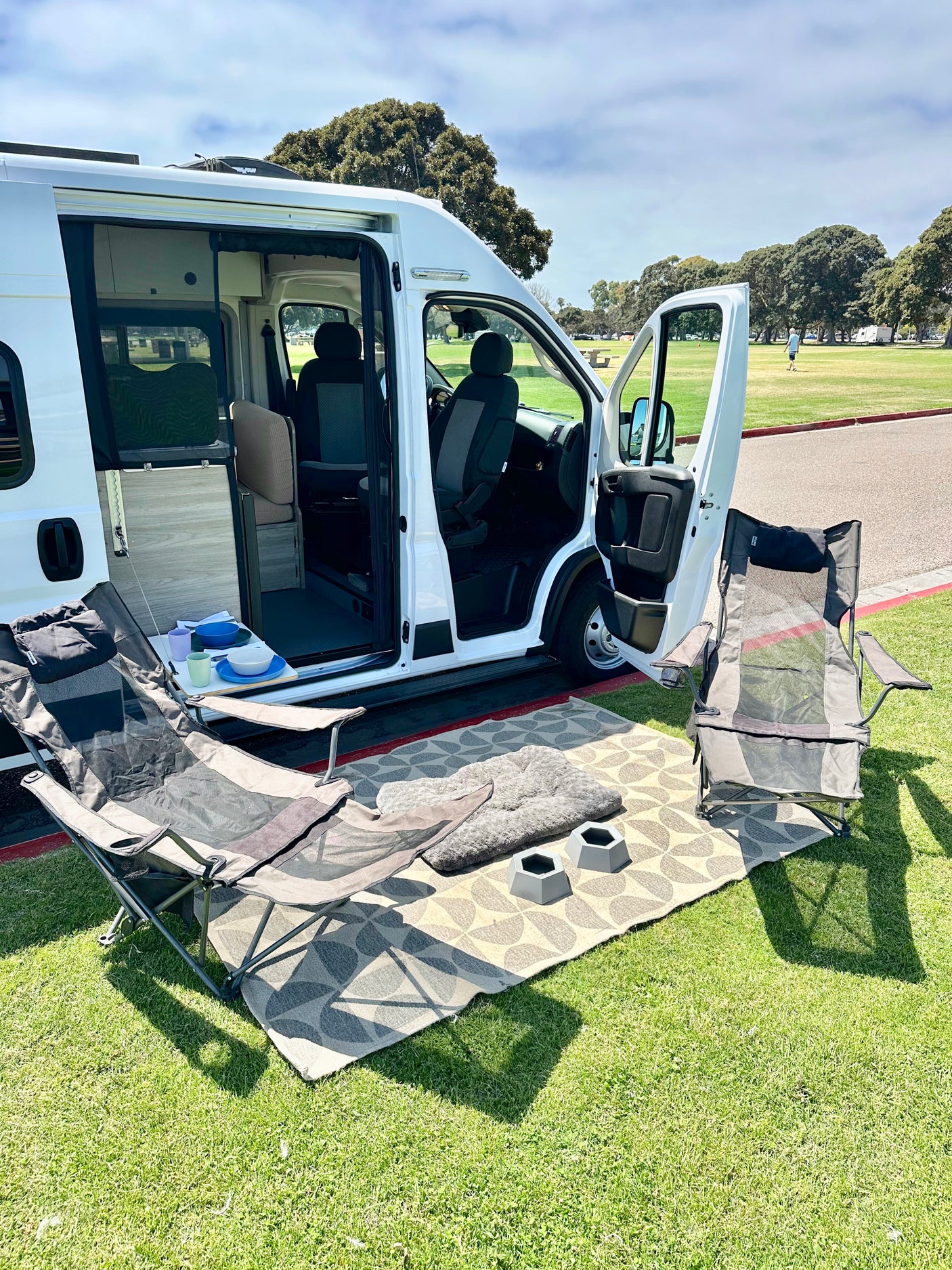 RV rental experience: White camper van open to a cozy outdoor setup on a rug with chairs, pet bed, and bowls in a park.