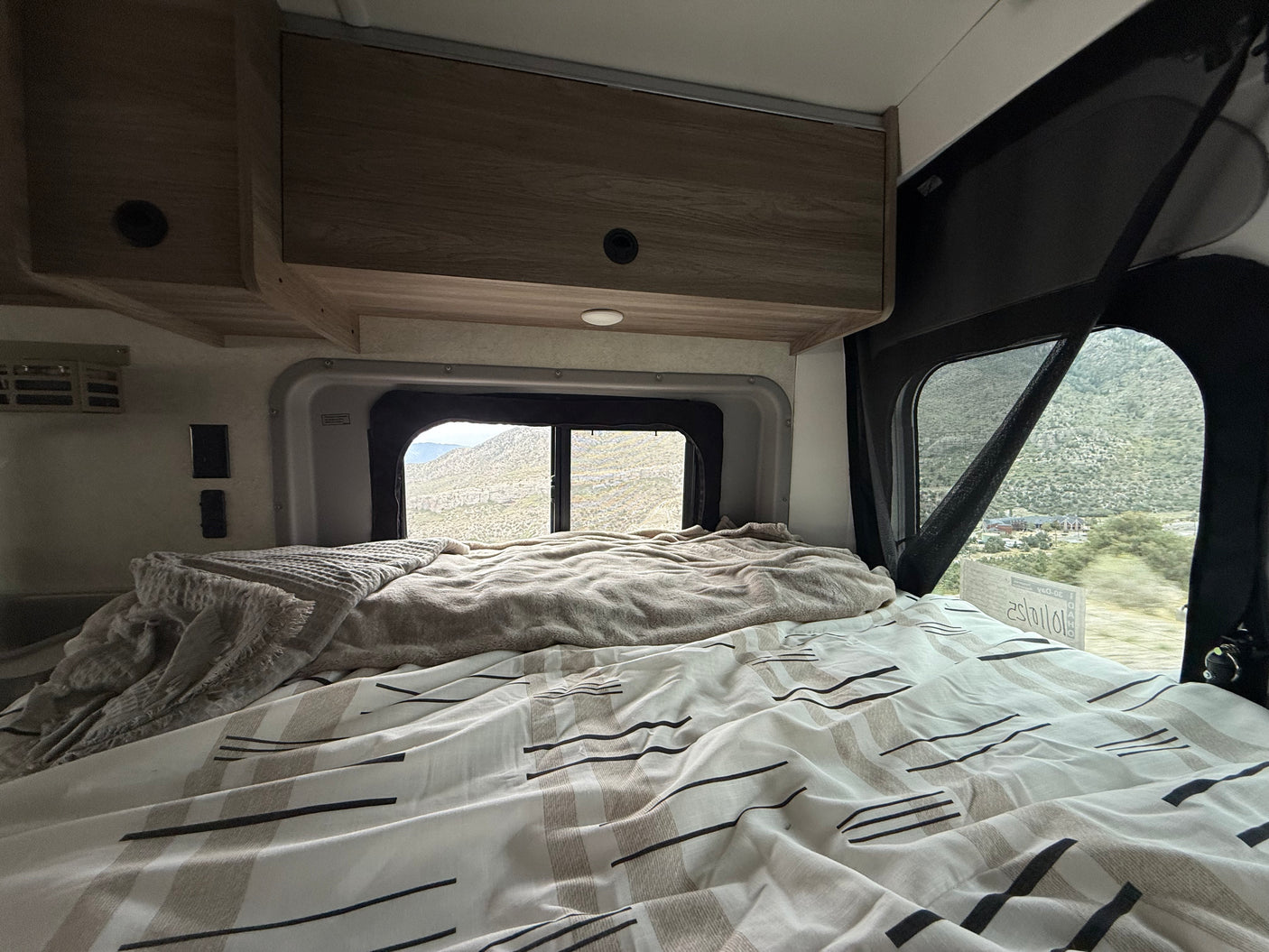 Cozy bed in an RV rental, offering scenic mountain views through the window. Overhead cabinets and reading light add comfort.